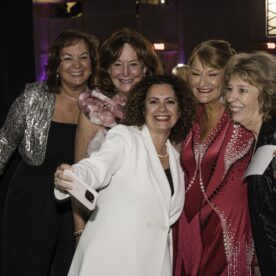 A group of five glamours women are dressed in black tie attire and taking a selfie.