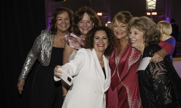 A group of five glamours women are dressed in black tie attire and taking a selfie.
